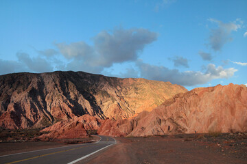 The asphalt highway across the desert at sunset. Beautiful panorama view of the empty road along the arid valley, the red sand, sandstone and rocky hills with a dusk light.