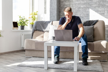 pensive adult man working at laptop from apartment while sitting on sofa