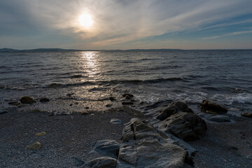 A sandy beach with some large rocks scattered throughout. The water has some small waves. The sky is setting and the sun is going down. There are clouds covering the sun leaving a ray of light.