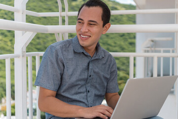 Young Latino shopping online using a laptop, sitting on a chair on his balcony