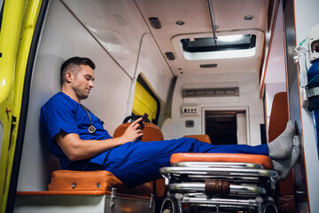 Male paramedic in a blue uniform resting and talking to someone over the video call on his smartphone in the ambulance car.