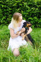 A beautiful blonde young woman sitting in green grass holds her cute dog in her arms. Vertical view