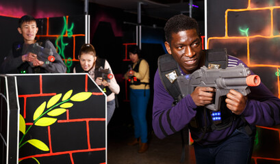 Fototapeta na wymiar Portrait of cheerful African American man aiming laser gun at other players during lasertag game in dark labyrinth