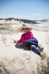 Naklejka na ściany i meble Young girl wearing a broad brim safe sun hat pink jumper jacket playing in the sand at the beach on a bright summer day 