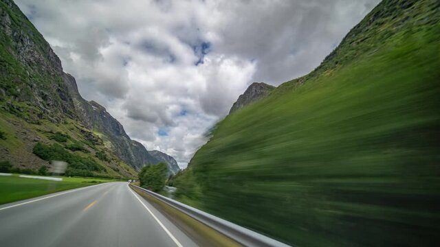 Drive on the E39 highway in Norway near Byrkjelo. The gray road going through the lush valley siding the river. Mountains fover on both sides of the road. Heavy, thick white-gray clouds flying above.
