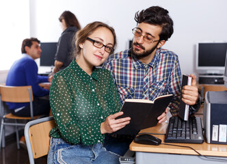 Couple of adult positive students reading textbook together while studying in university computer class