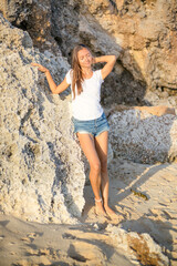 Slim young woman standing barefoot on the sand near the rock. Caucasian woman wearing jeans shorts, white T-shirt. Enjoying sunlight at the beach. Vacation in Asia. Travel lifestyle. Bali