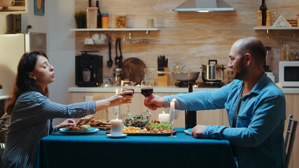 Young caucasian couple drinking a glass of wine during romantic dinner. Relax happy people clinking, sitting at table in kitchen, enjoying the meal, celebrating anniversary in the dining room