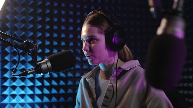 Beautiful young girl dj speaks into microphone, records podcast or morning news show for radio. Close up of podcast presenter or DJ talking on the microphone. Wears professional headphones