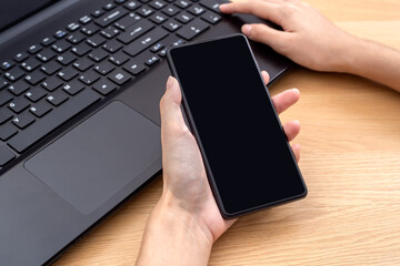 Female hand holding mobile phone with blank screen over laptop.