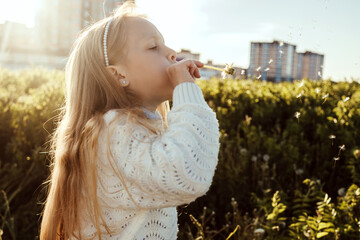 A girl blows on a dandelion in a field. Houses on background.