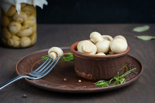 Marinated or fermented champignon mushrooms served in clay bowl with fork on plate against glass can or jar on dark wooden background with bay leaf, dill and pepper at kitchen. Image with copy space