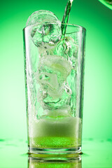 pouring green tarragon lemonade into glass with pieces of pure real ice on green gradient background