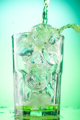pouring green tarragon lemonade into glass with pieces of pure real ice on green gradient background