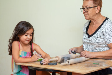A Senior Woman Is Teaching A Little Girl How To Form With Black Clay. Grandmother Helping Beautiful Grandchild Sculpt Dough Figurines In The Home.