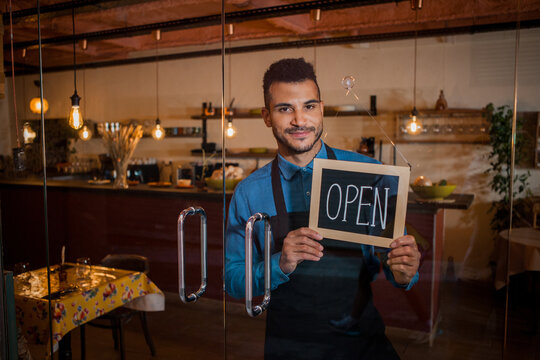 Attractive smiling waiter holding chalkboard with open inscription in cafe. Young african american entrepreneur leaning at the cafeteria door and looking at camera. Small business concept, restaurant