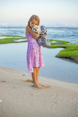 Cute little girl holding small white chihuahua dog with pink tail on the beach. Take care of the pet. Dog covered with scarf. Spend time outdoors. Bali, Indonesia