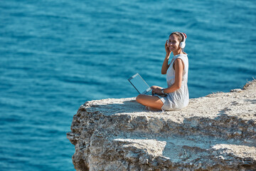 Happy lady working on laptop from trip. Smiling woman in headset. Ocean view and nature. Freelance job and travel idea