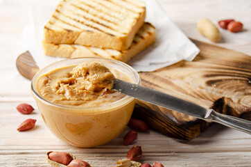Low angle view at glass bowl with peanut butter on white wooden table with toasts aside. Healthy...