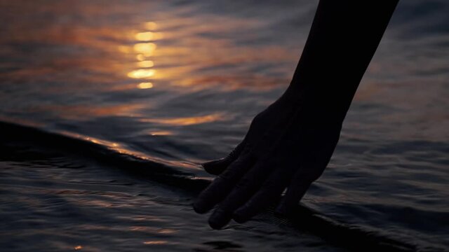 Woman scoops sea water against the sunset. Close-up hand touches the surface of the water.