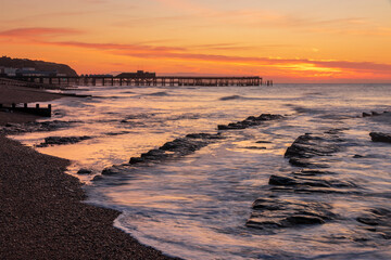 The sun rising behind Hastings pier in east Sussex and the tide receding on St Leonards beach revealing the submerged rocks