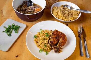 Dining table with baked chicken leg and spelled wheat in plate and pot.