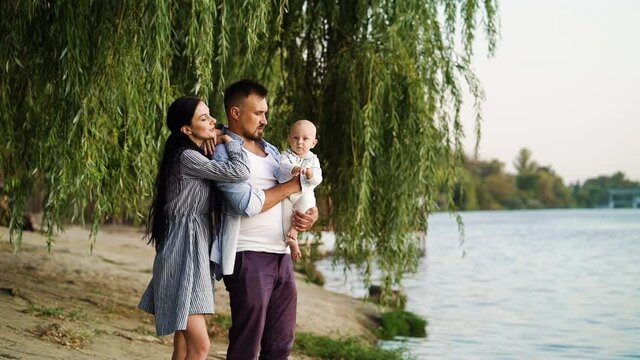 Young parents with baby standing under willow tree near river. Loving family spending time together in nature. Concept of bonding