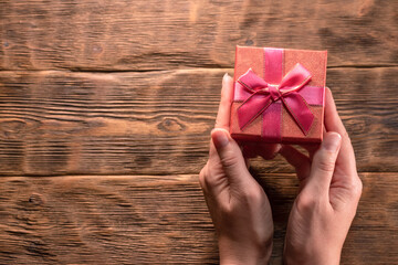 A small red gift box in female hands on the wooden table background with copy space.