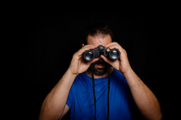 Bearded man dressed in blue t-shirt with binoculars posing against black background
