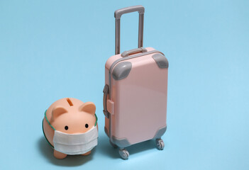 Travel during covid-19. Mini plastic travel suitcase and piggy bank with face mask on blue background.