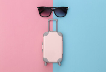 Travel concept. Mini plastic travel suitcase and sunglasses on pink blue pastel background. Minimal...