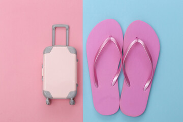 Travel or beach resort flat lay. Mini plastic travel suitcase and flip flops on pink blue pastel background. Minimal style. Top view