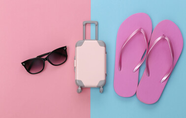 Obraz na płótnie Canvas Travel or beach resort flat lay. Mini plastic travel suitcase and flip flops, sunglasses on pink blue pastel background. Minimal style. Top view
