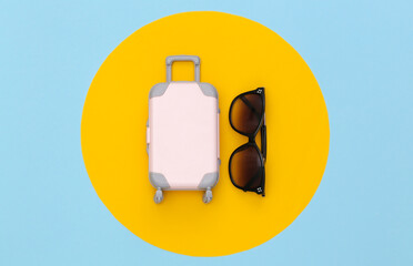 Travel concept. Mini plastic travel suitcase and sunglasses on blue pastel background with yellow circle. Minimal style. Top view, flat lay