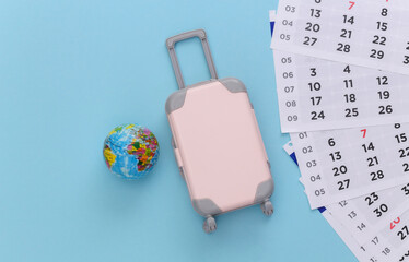 Time to travel. Mini plastic travel suitcase, globe and monthly calendar on blue background. Minimal style. Top view. Flat lay