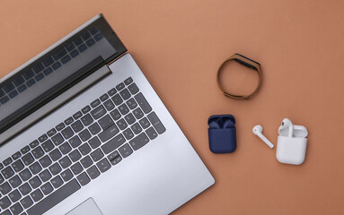 Laptop and wireless headphones with charger case and smart bracelet on brown background. Top view