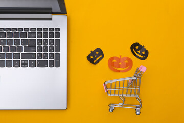 Laptop and shopping trolley with paper cut halloween pumpkins on a yellow background. Halloween theme. Top view - Powered by Adobe