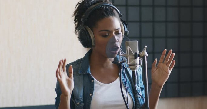 Young black woman recording new song in modern sound studio, singing in microphone and dancing