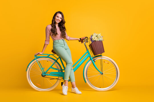 Full length body size photo of pretty girl sitting on bicycle with basket of flowers smiling isolated on vivid yellow color background