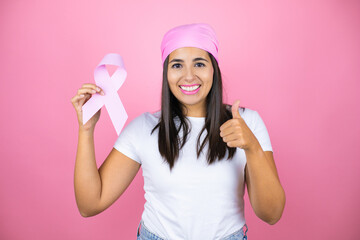 Obraz na płótnie Canvas Young beautiful woman wearing pink headscarf holding brest cancer ribbon over isolated pink background