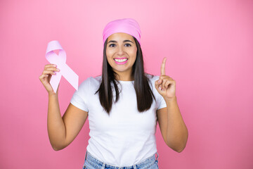 Young beautiful woman wearing pink headscarf holding brest cancer ribbon over isolated pink background surprised with an idea or question pointing finger with happy face, number one