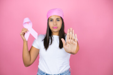 Obraz na płótnie Canvas Young beautiful woman wearing pink headscarf holding brest cancer ribbon over isolated pink background with open hand doing stop sign with serious and confident expression, defense gesture