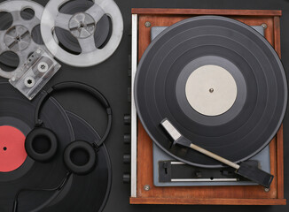 Retro vinyl record player with records, audio magnetic reel, audio cassette and stereo headphones on black background. Top view. Flat lay