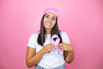 Young beautiful woman wearing pink headscarf holding brest cancer ribbon over isolated pink background smiling and putting the ribbon in the heart