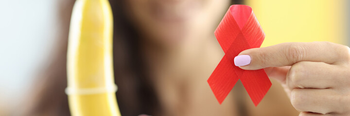 Woman holds in her hands banana, which is wearing condom and red ribbon. HIV infection and...