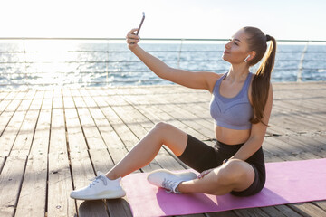 Slm fit woman in sportswear makes selfie on a smartphone while sitting on the mat on the beach at sunrise