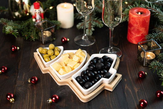 Christmas tree plate with green and black olives, cheese. Glasses of champagne, nutcracker and fir tree branches on old wooden background, dark image in rustic style, selective focus