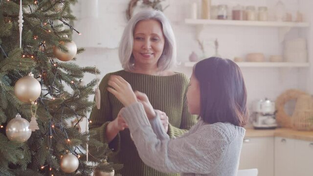 Little Asian girl decorating Christmas tree at home with assistance of beautiful grandmother while preparing home for holiday