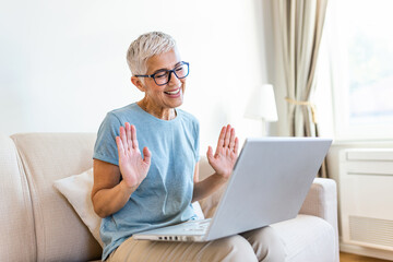 Happy mature woman waving to someone while having a video call over laptop at home. Gray-haired senior woman waving hand in front of laptop while having video call with her family members.