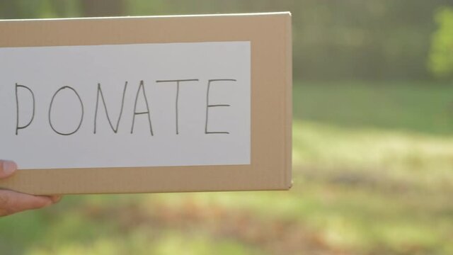A man brings into the frame a paper box with the words "donate" on a background of green grass.